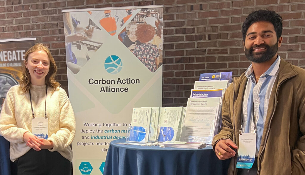 CAA staff Diana Leane stands in a white sweater and black pants next to a Carbon Action Alliance banner and table with Vilas Annavarapu standing to the right in a brown jacket and brown slacks.